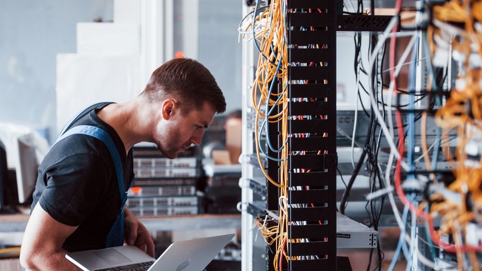 Young man in uniform and with laptop works with internet equipment and wires in server roomYoung man in uniform and with laptop works with internet equipment and wires in server room.server, network, technician, laptop, room, computer, cable, engineer, system, data, man, guy, male, support, service, information, administrator, work, job, occupation, rack, industry, center, technology, science, connection, research, cabinet, indoors, repairing, manager, fixing, router, solution, security, caucasian, web, digital, lan, one, person, equipment, storage, installation, operating, electric, professional, uniform, ethernet, internet