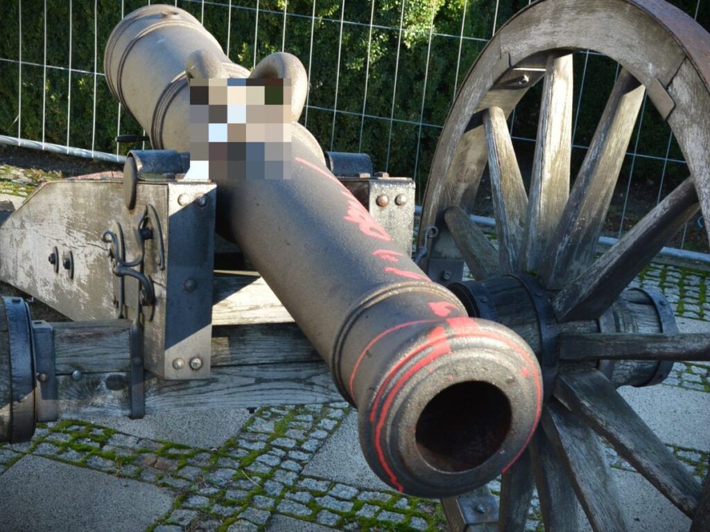 Działo na drewnianym wózku, z widocznymi śladami rdzy, ustawione na brukowanej nawierzchni w otoczeniu zieleni.