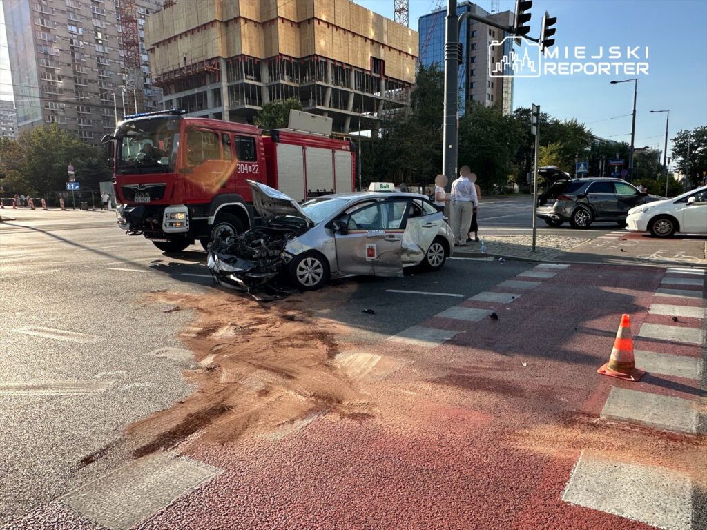 Zniszczony samochód osobowy stoi na skrzyżowaniu, obok wozu straży pożarnej, a kierowcy rozmawiają na chodniku.