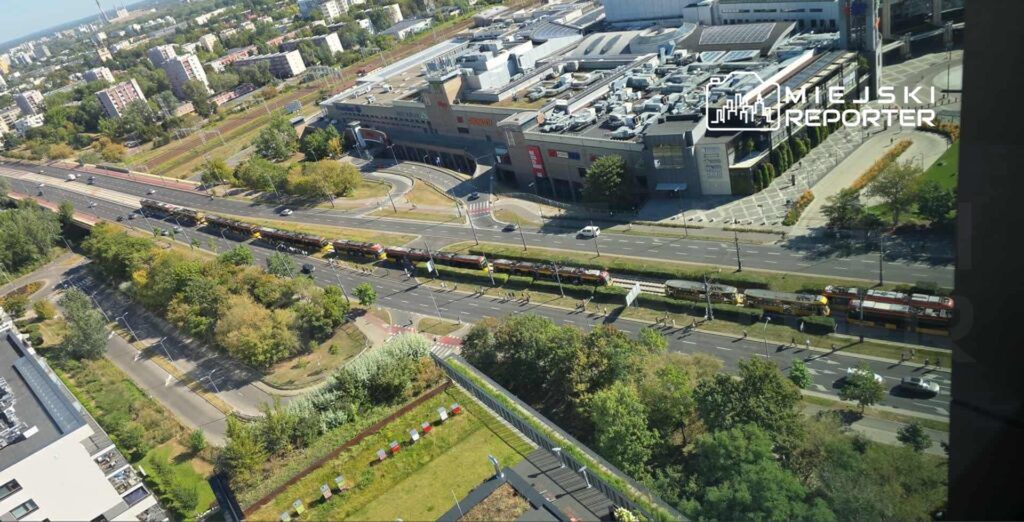 Widok z góry na ruchliwą ulicę z tramwajami, obok nowoczesnego centrum handlowego otoczonego zielenią.
