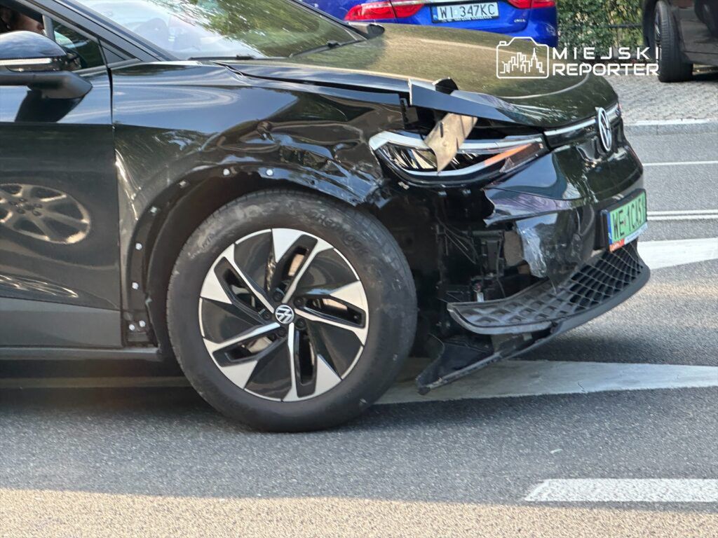 Czarny samochód elektryczny z uszkodzoną przednią częścią stoi na ulicy, obok innych pojazdów.