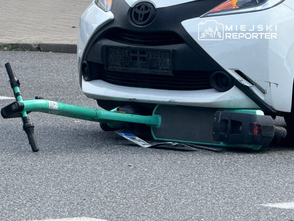 Zielona hulajnoga leży pod białym samochodem osobowym na asfaltowej nawierzchni drogi.