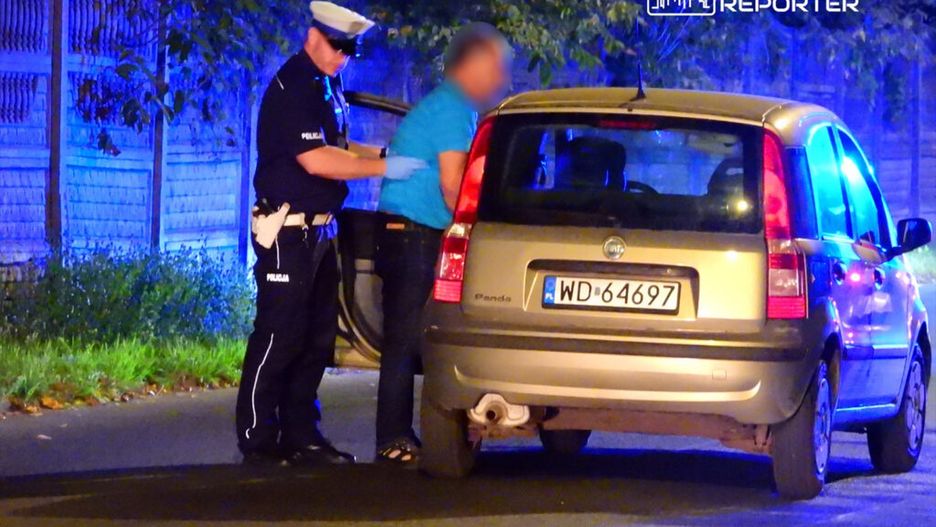 Policjant w mundurze kontroluje mężczyznę przy otwartym bagażniku samochodu osobowego w nocy.