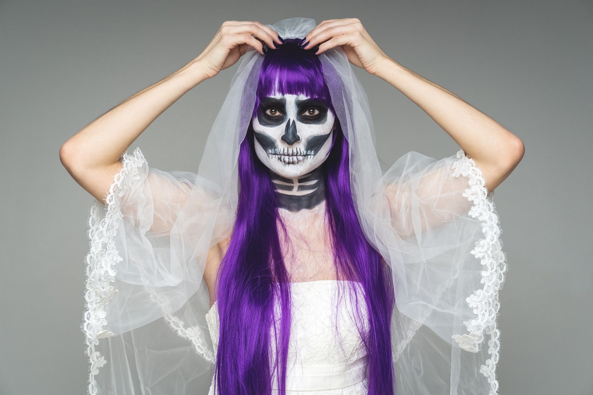 Portrait of woman looks at the camera with terrifying halloween skeleton makeup and purple wig corrects bridal veil, wedding dress over gray background. Black wedding. Portrait of woman looks at the camera with terrifying halloween skeleton makeup and purple wig corrects bridal veil, wedding dress over gray background. Black wedding. black bride, black wedding, bride, calavera, calaverita, catrina, celebration, copy space, day, day of dead, dead, dead bride, death, devil, dia de muertos, evil, face, girl, gothic, hair, halloween, horror, katrina, make up, makeup, mask, mexican, mexico, moke up, newlywed, paint, purple, purple hair, saints, scary, skeleton, skull, skull sugar, spooky, sugar, traditional, voodoo, wedding, wedding dress, witch, waiting for the bride, black bride, black wedding, bride, calavera, calaverita, catrina, celebration, copy space, day, day of dead, dead, dead bride, death, devil, dia de muertos, evil, face, girl, gothic, hair, halloween, horror, katrina, make up, makeup, mask, mexican, mexico, moke up, newlywed, paint, purple, purple hair, saints, scary, skeleton, skull, skull sugar, spooky, sugar, traditional, voodoo, wedding, wedding dress, witch, waiting for the bride