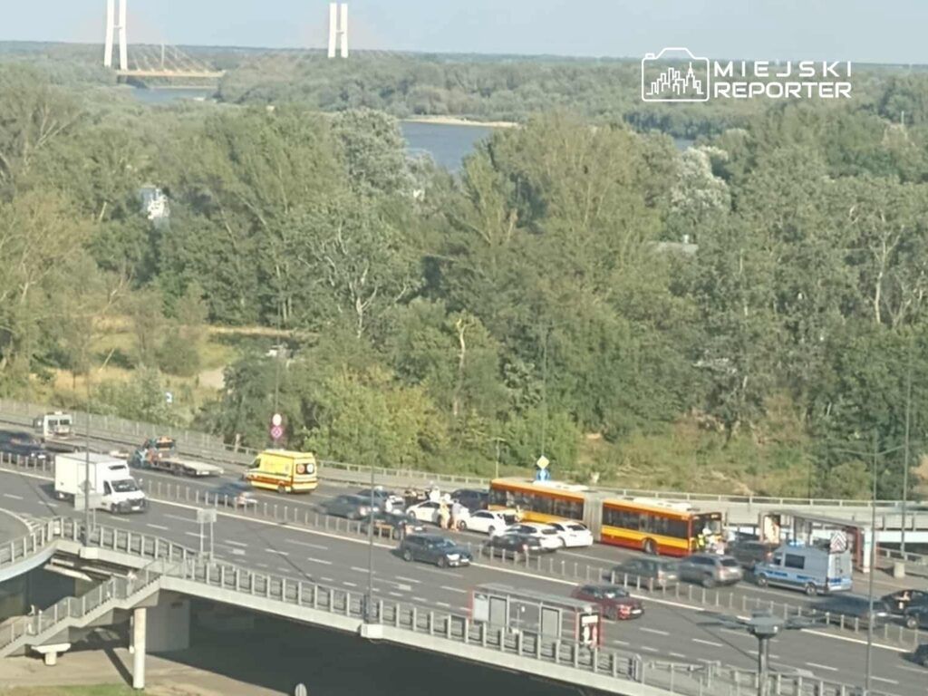 Na zdjęciu widoczny jest ruch na drodze w pobliżu mostu, z pojazdami w różnych kierunkach. W centralnej części znajduje się karetka pogotowia oraz kilka samochodów osobowych i autobusów. W tle widać zieleń oraz rzekę. Obraz ilustruje sytuację drogową w mieście.