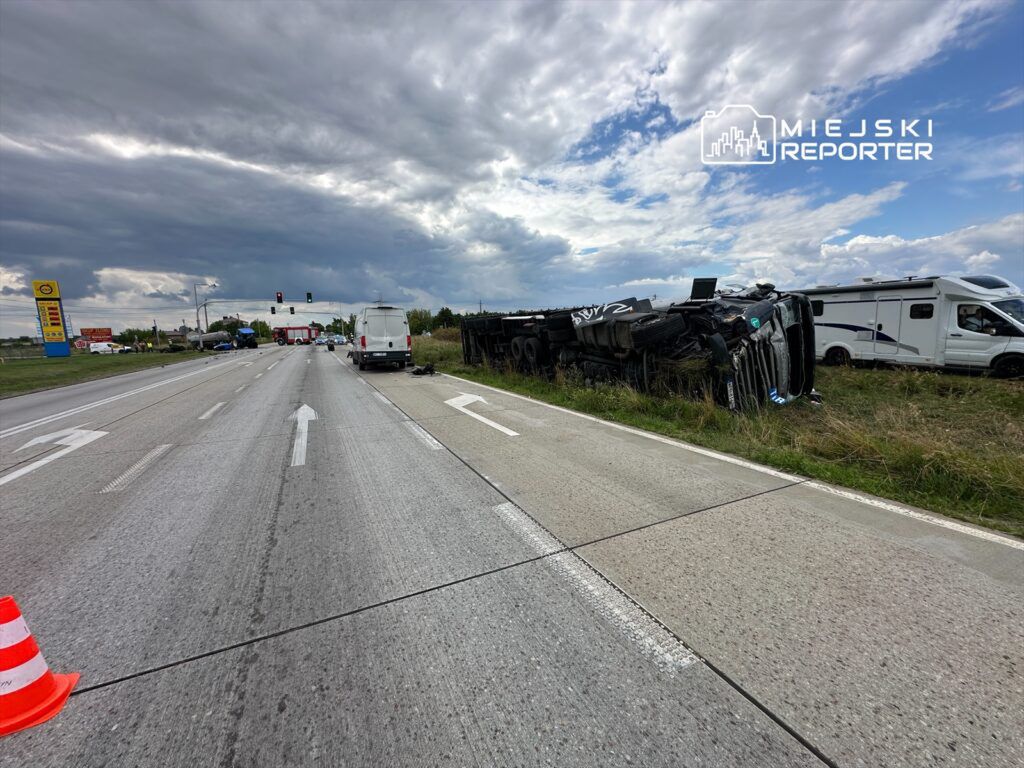 Na drodze leży przewrócony ciężarówka, obok stoją pojazdy i służby ratunkowe. W tle widoczne są chmury.