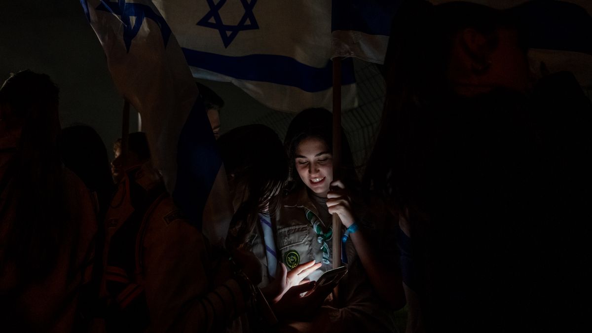 PETAH TIKVA, ISRAEL - NOVEMBER 26: People look at their phones while waving Israeli flags ahead of the helicopter enroute with Israeli hostages released earlier by Hamas lands at Schneider medical centre on the third day of the temporary truce where they disembarked behind screens as family and friends wait nearby on November 26, 2023 in Petah Tikva, Israel. A temporary truce between Israel and Hamas began on Friday, followed by the return of some Hamas-held hostages as well as the release of Palestinian prisoners. If the truce holds, around 50 Israeli hostages are to be released, among the approximate 240 that Israeli authorities say Hamas took captive during its Oct. 7 attack. (Photo by Alexi J. Rosenfeld/Getty Images)