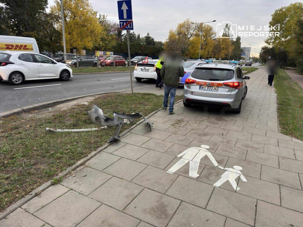 Uszkodzona część samochodowa leży na trawniku obok chodnika, obok stojących pojazdów i znaków drogowych.