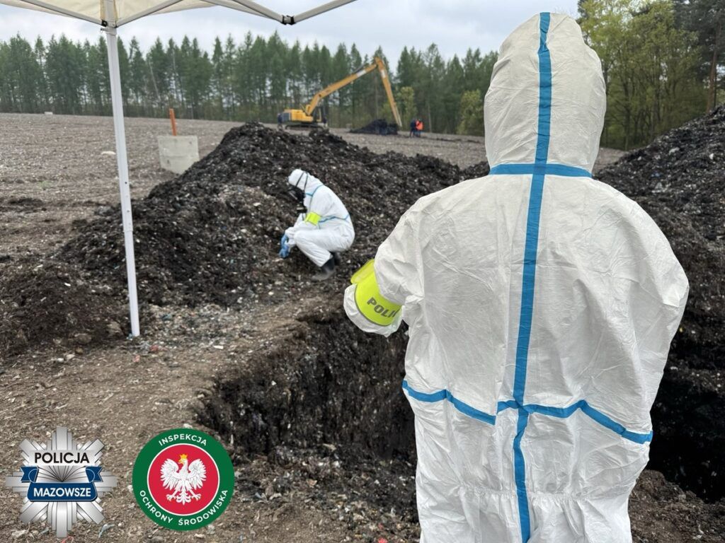 Funkcjonariusze w kombinezonach ochronnych przeszukują teren z odpadami, w tle widoczny koparka.