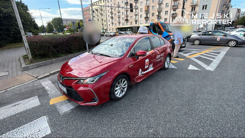 Zdarzenie drogowe z udziałem taksówki Uber na ulicy w mieście. Na zdjęciu widoczny uszkodzony czerwony samochód osobowy oraz osoby zajmujące się sytuacją. Tło przedstawia miejską architekturę oraz inne pojazdy na drodze. Służby porządkowe są w trakcie interwencji.