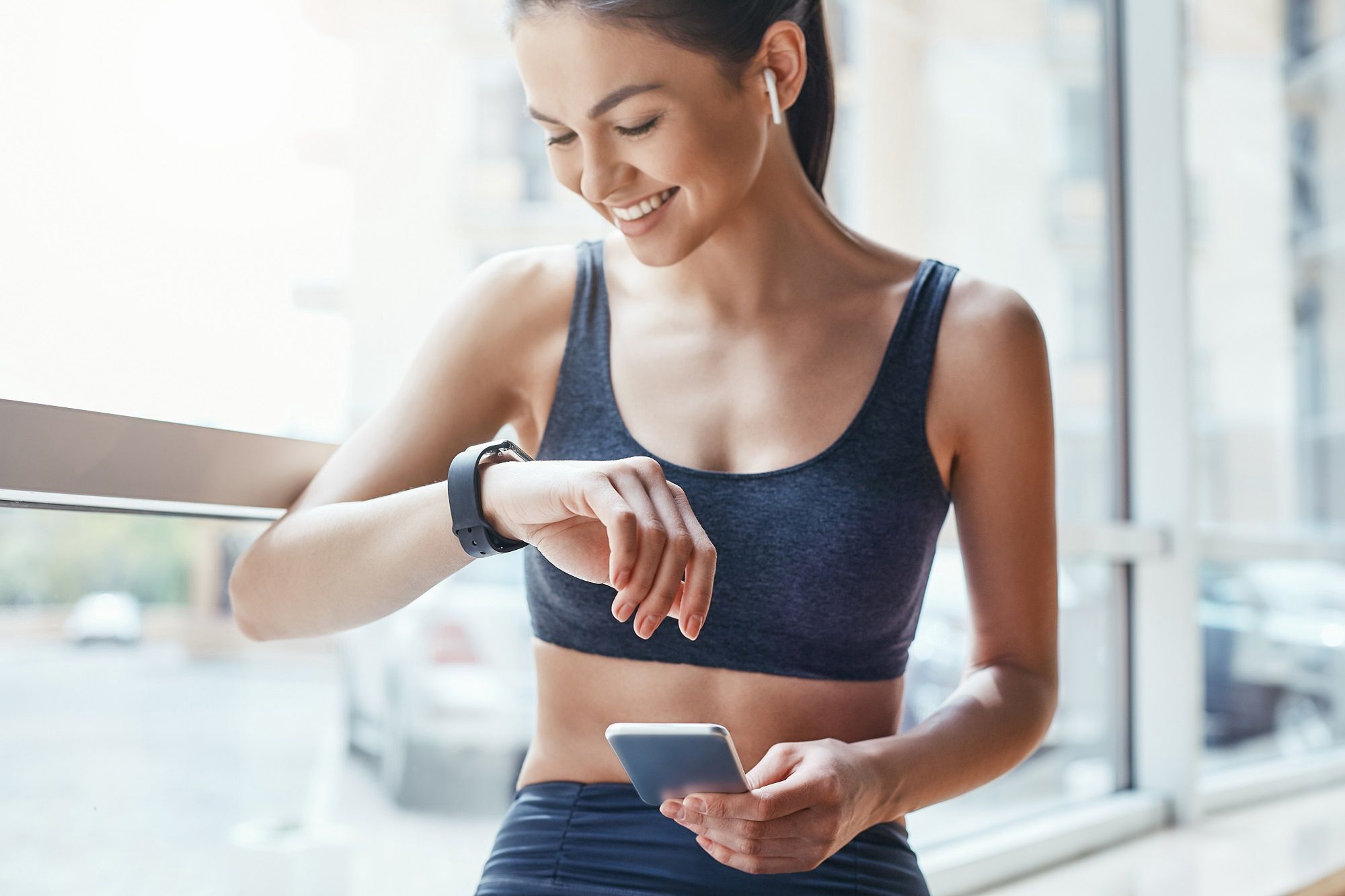 Making training easier. Young and cheerful woman in sportwear looking at her sport bracelet and using smartphone while standing in front of window at gymMaking training easier. Young and cheerful woman in sportwear looking at her sport bracelet and using smartphone while standing in front of window at gym. Fitness concept. Sport and healthy lifestyle. Technologiesadult, athlete, attractive, beautiful, beauty, body, bra, built, caucasian, checking, checking the time, close up, clothing, club, concentration, exercising, exhaustion, females, gym, headphones, healthy, healthy lifestyle, human hand, lifestyle, motivation, muscle, music, new approach, one person, one woman only, outdoors, pedometer, phone, professional sport, relaxation, smart, smart watch, sport, sports clothing, sports person, sports training, time, training, watch, wellbeing, woman, women, young adult, app, smile