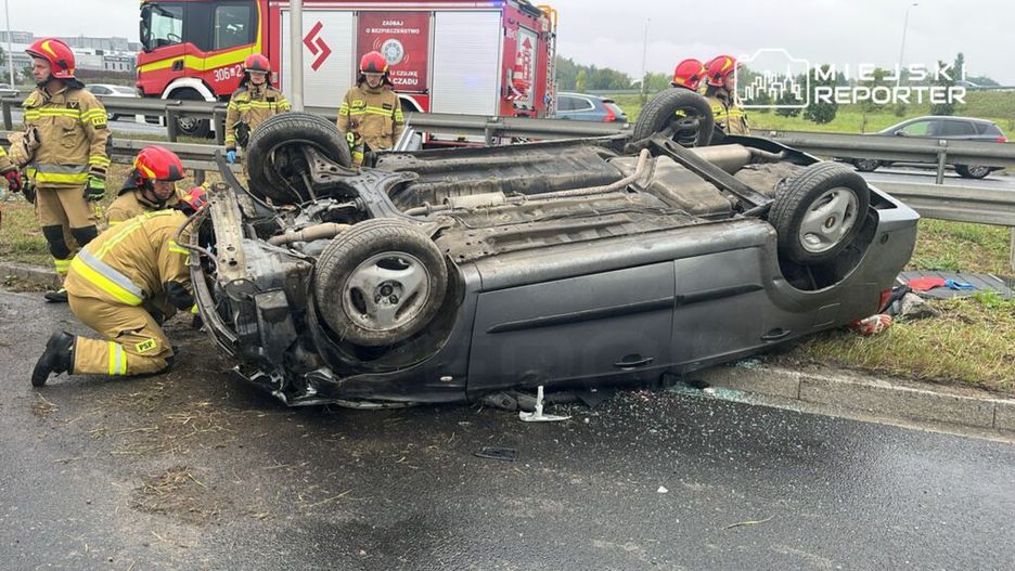 Strażacy w mundurach zabezpieczają przewrócone na dach auto na poboczu drogi, obok widać pojazd strażacki.