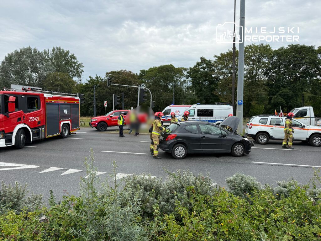 Na skrzyżowaniu zatrzymane pojazdy ratunkowe, w tym wóz strażacki i ambulans, podczas akcji ratunkowej po wypadku drogowym.