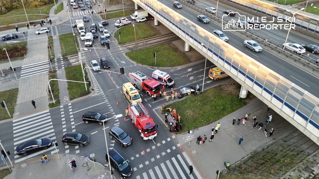 Na skrzyżowaniu ulicy ruch samochodowy jest wstrzymany z powodu wypadku, w którym przewrócił się samochód. Ratownicy i strażacy udzielają pomocy poszkodowanym, a obok stoją pojazdy służb ratunkowych.