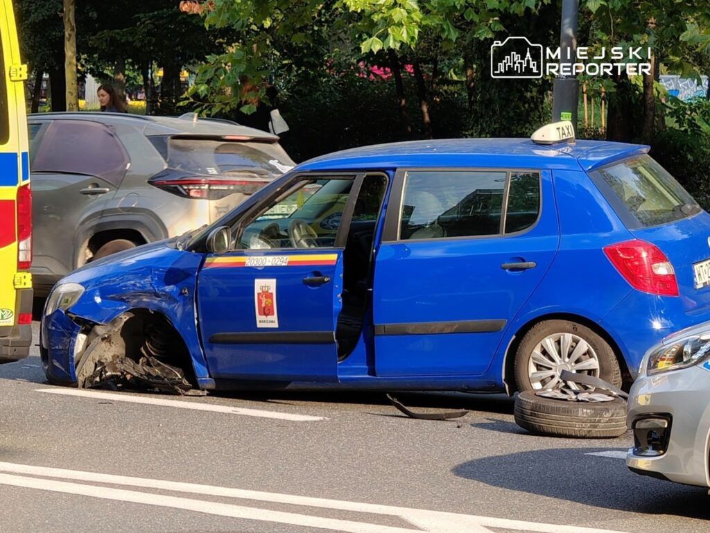 Niebieskie taxi z uszkodzoną oponą zatrzymane na jezdni, obok stoją samochody i ambulans.