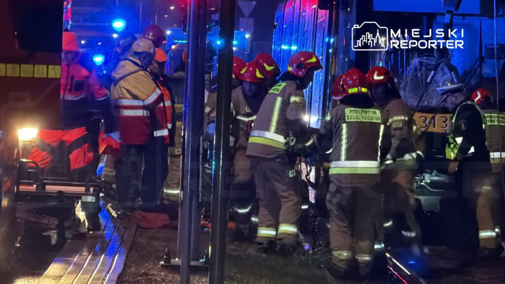 Grupa strażaków w kaskach i mundurach udziela pomocy poszkodowanym przy zderzeniu tramwaju w nocy, otoczeni niebieskimi światłami.