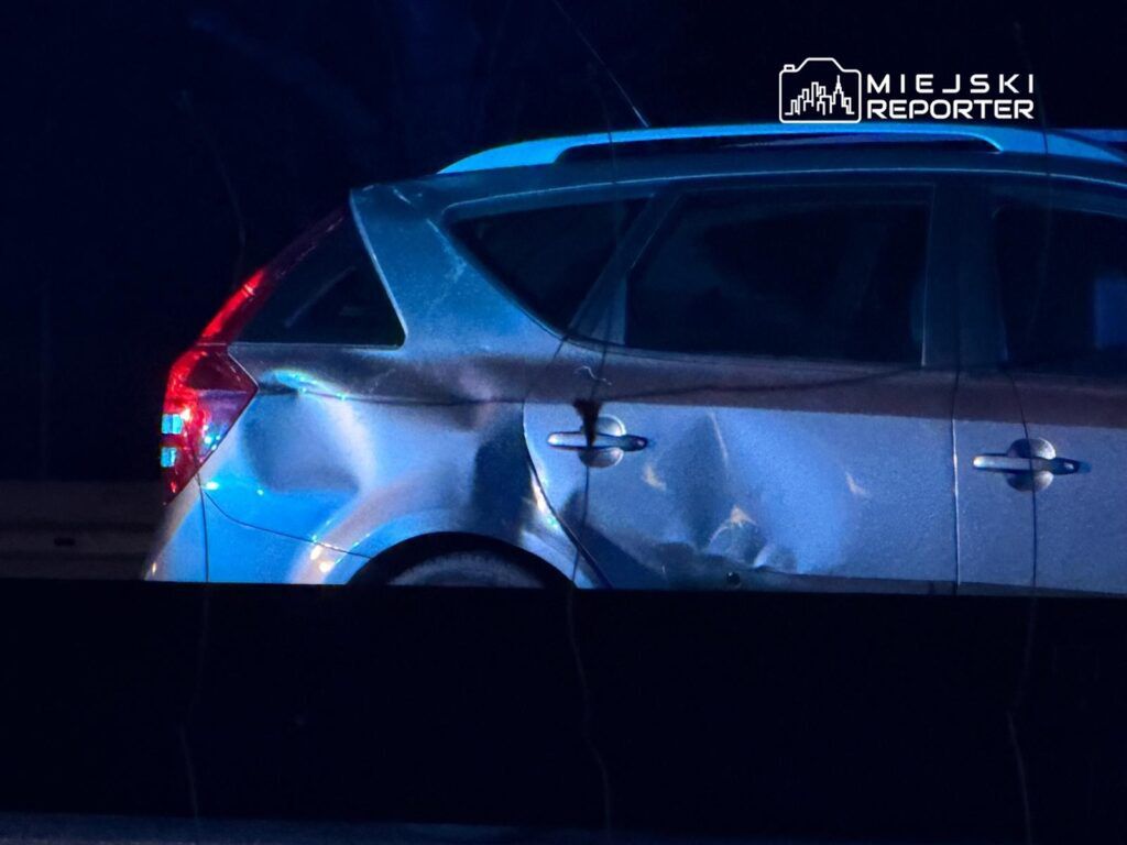 Uszkodzony samochód osobowy z wgnieceniem na drzwiach, zaparkowany w ciemnym otoczeniu.