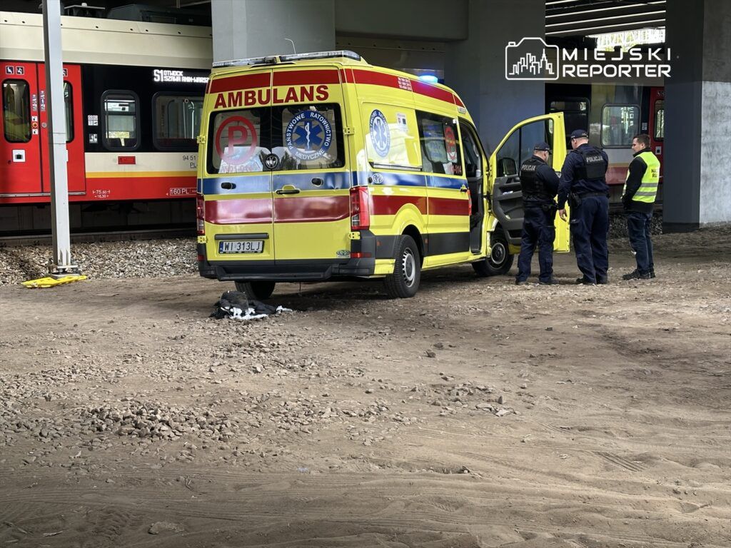 Zespół ratunkowy w żółtej karetce omawia sytuację z policjantami pod torami kolejowymi.