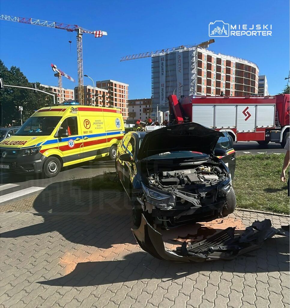 Czarny samochód osobowy z uszkodzoną maską stoi na trawniku obok karetki pogotowia i wozu strażackiego.