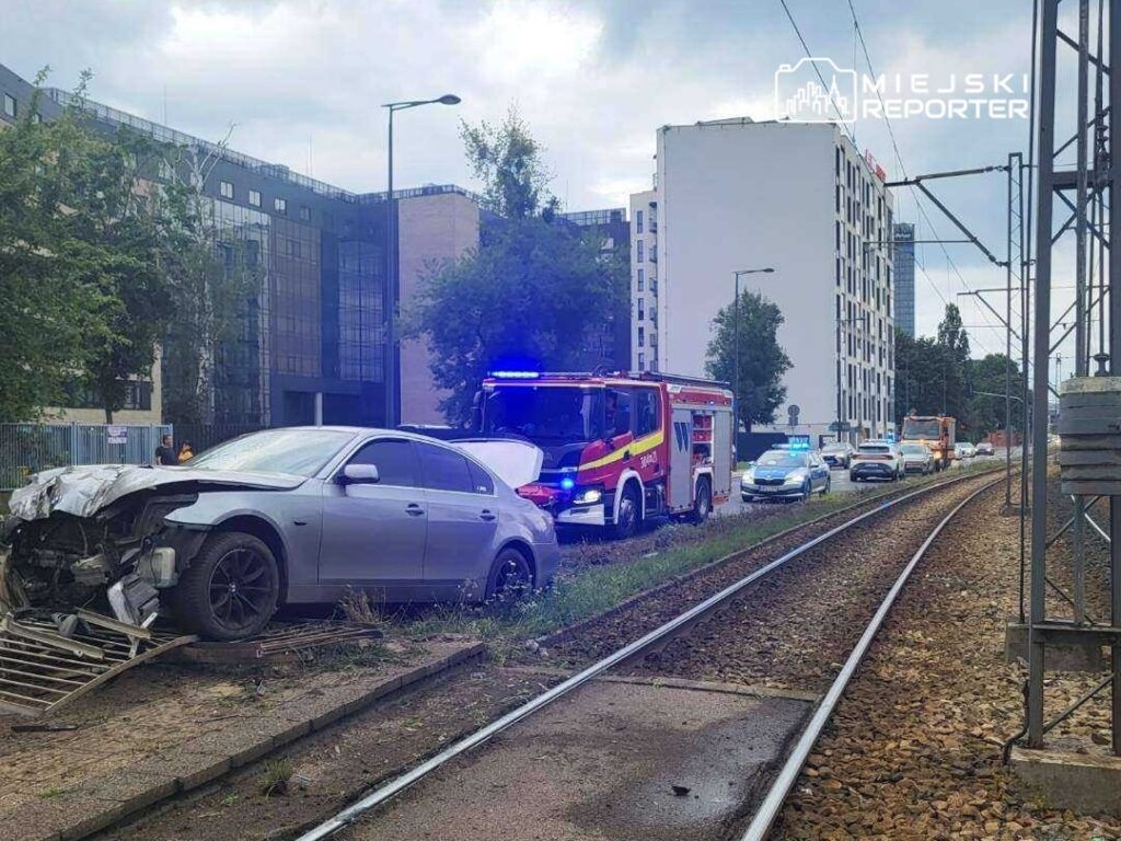Uszkodzony samochód osobowy stoi na poboczu torów kolejowych, obok zjeżdżają pojazdy służb ratunkowych.