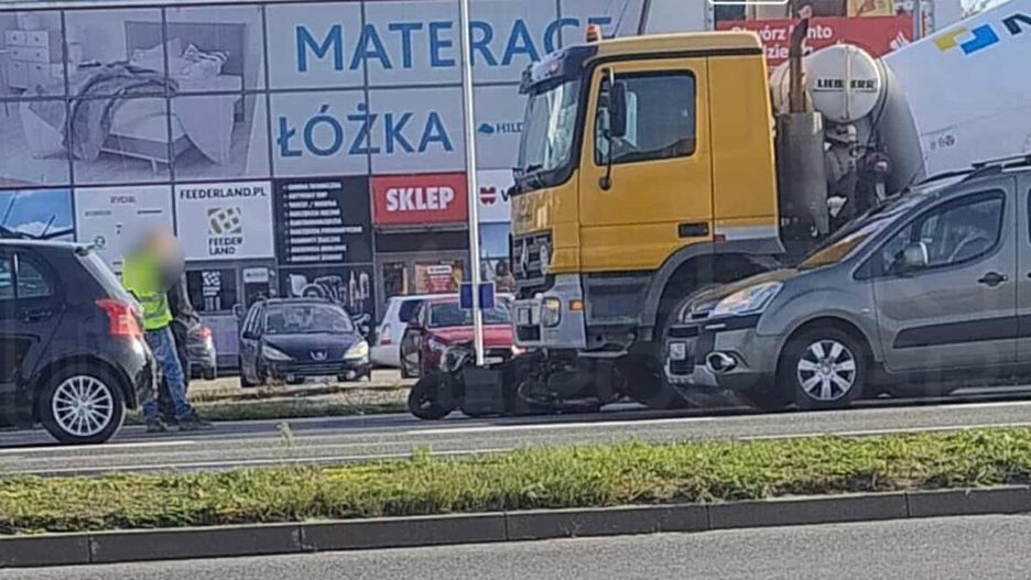 Mężczyzna w kamizelce odblaskowej rozmawia z kierowcą na ruchliwej ulicy obok dużego billboardu reklamowego.