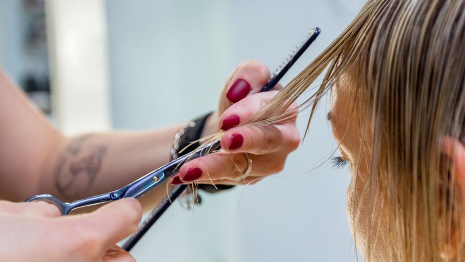 Hands of a professional hairdresser scissors the bangs of wet hair of a blonde woman. Fashionable hairstyle design in a beauty salon. Small female business