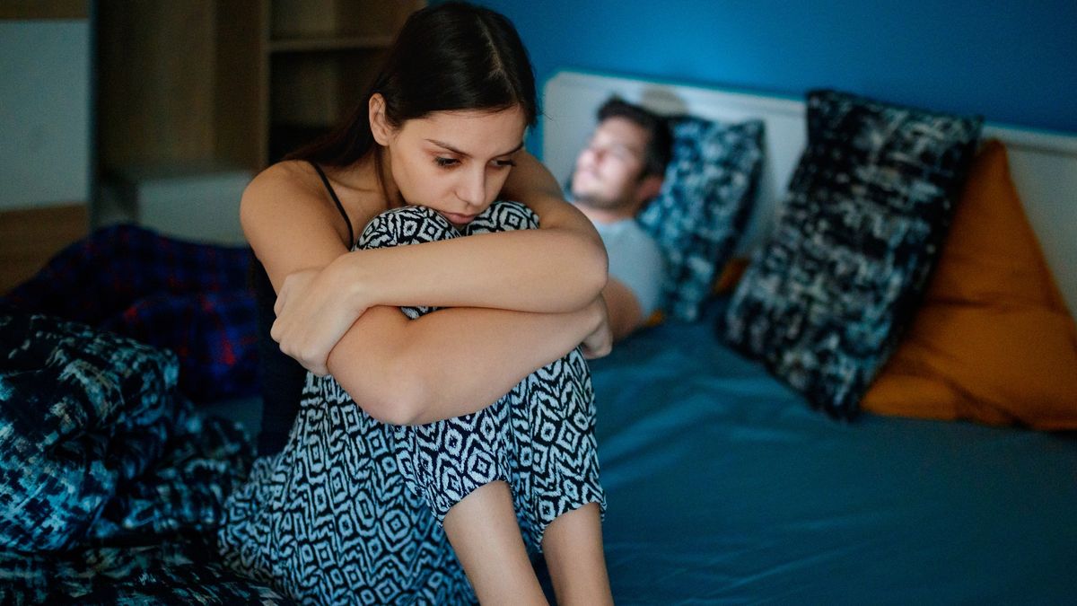 Sad woman sitting on bed with partner in background
Sad girlfriend sitting on bed edge, holding her legs after fight, her partner is lying in bed in background, his face is lit by smart phone
zoranm