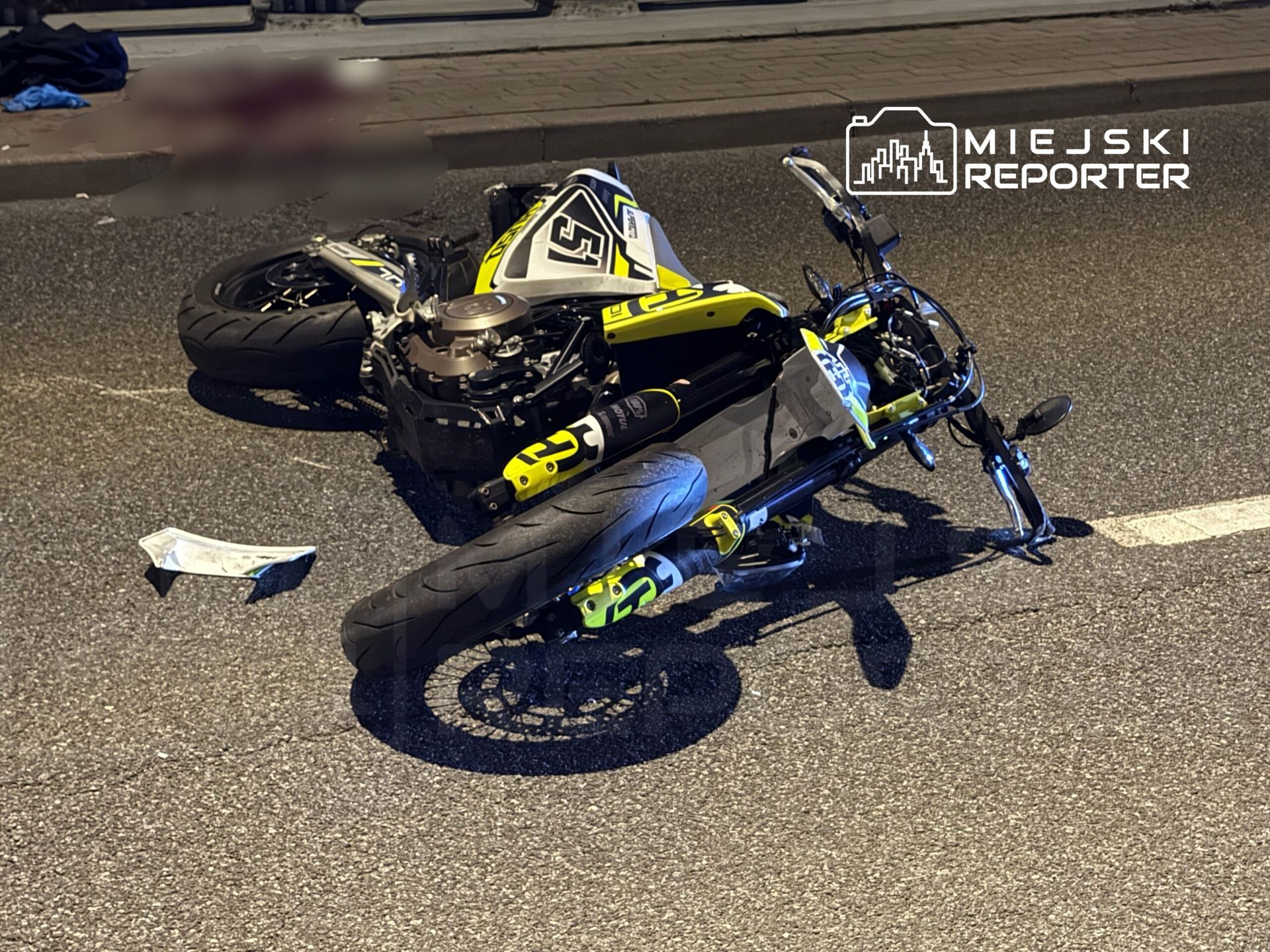Motocykl leżący na jezdni z uszkodzoną obudową, w tle fragmenty odzieży na chodniku.