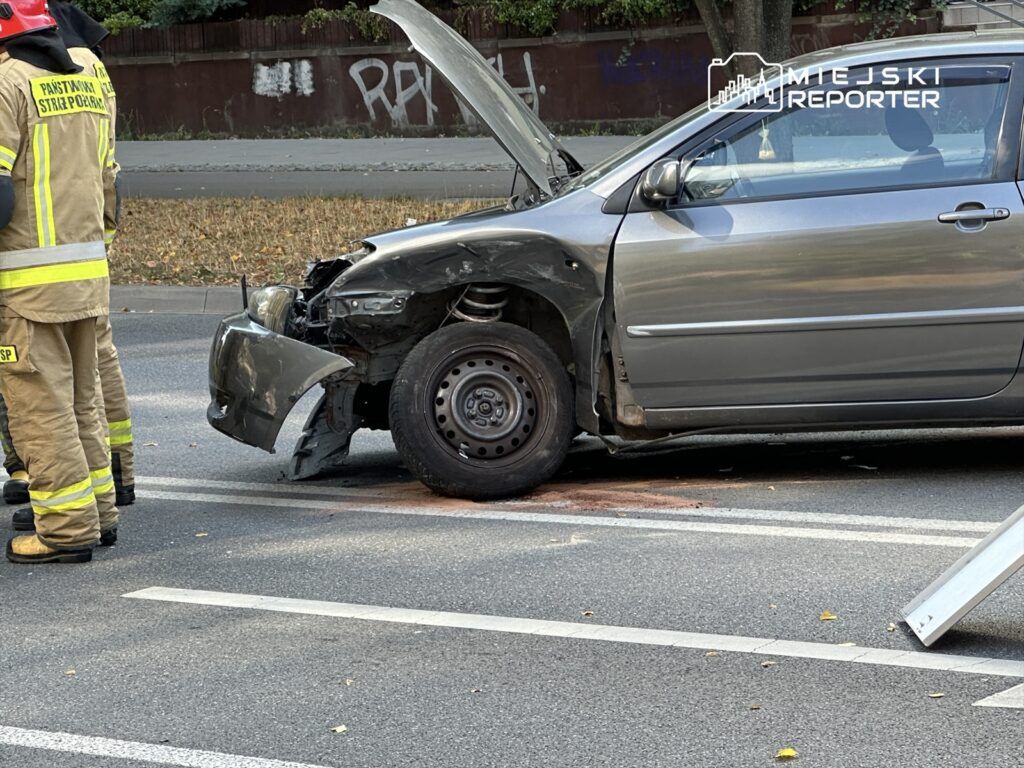 Strażacy w mundurach rozmawiają obok uszkodzonego samochodu z otwartą maską na ulicy.