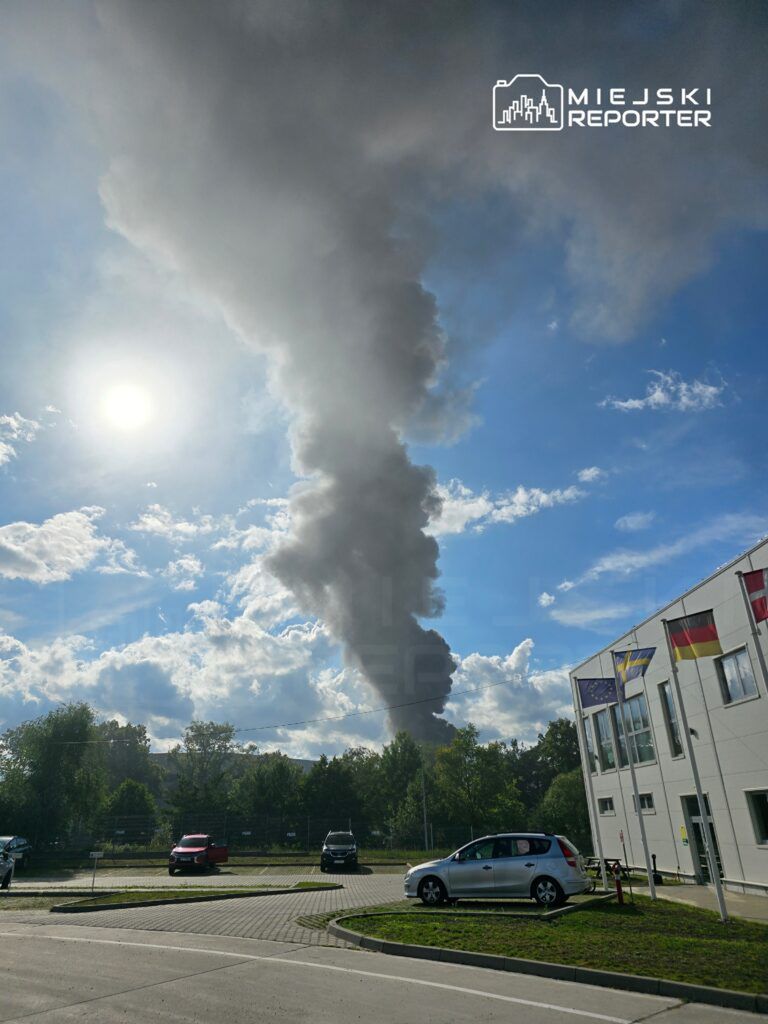 Widok na niebo z dużą chmurą dymu unoszącą się z horyzontu. W tle widać budynki oraz flagi. Słońce świeci na tle chmur, co tworzy kontrast z ciemnym dymem. Obraz ilustruje sytuację, która może być związana z pożarem lub innym zdarzeniem emitującym dym.