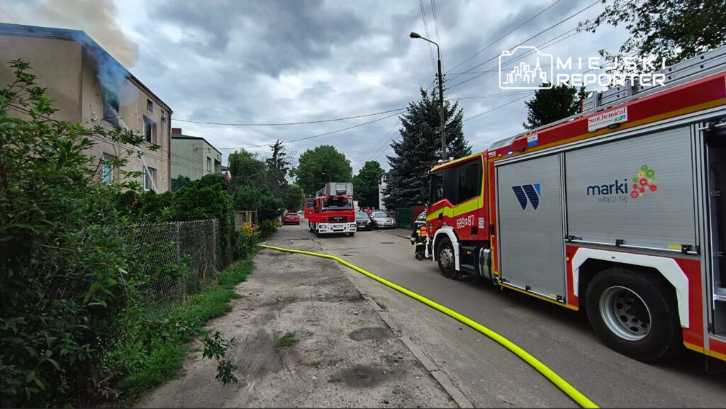 W tle dym wydobywający się z okna budynku, strażacy przy wozach gaśniczych rozkładają węże wzdłuż ulicy.