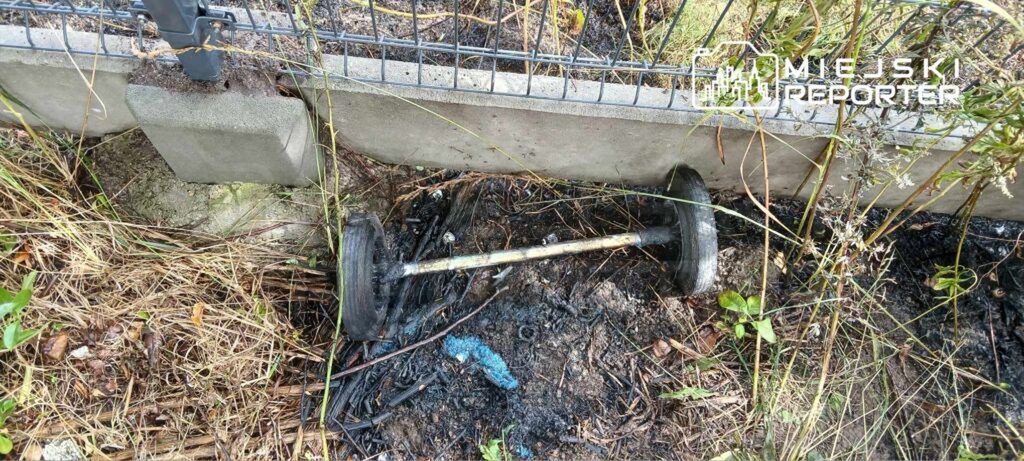 W ogrodzie leży uszkodzona sztanga obok spalonej trawy i zarośli, w pobliżu betonowego ogrodzenia.