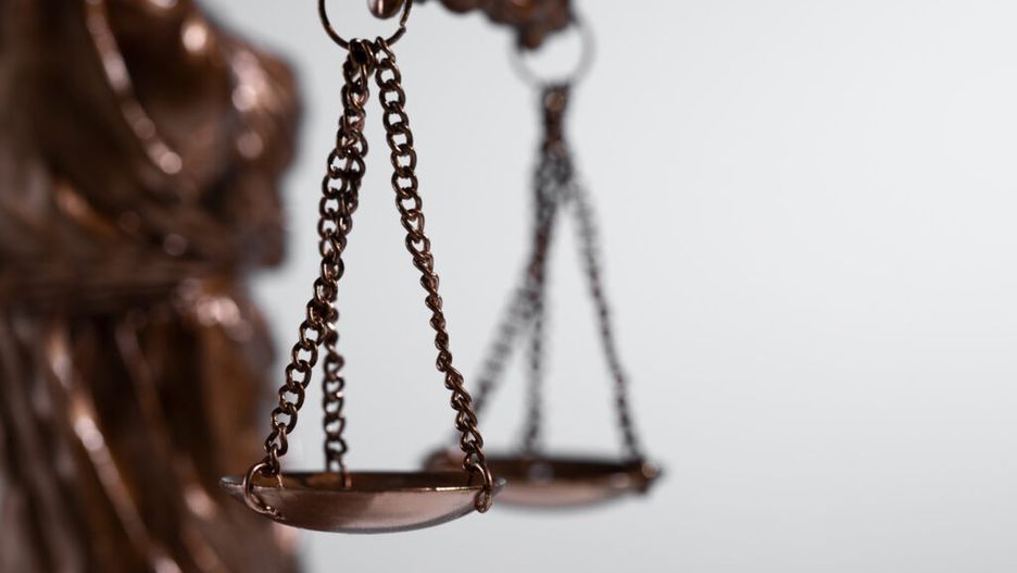 Waga sprawiedliwości w brązowym kolorze zawieszona na łańcuszkach, w tle rozmyta figura symbolizująca prawo.