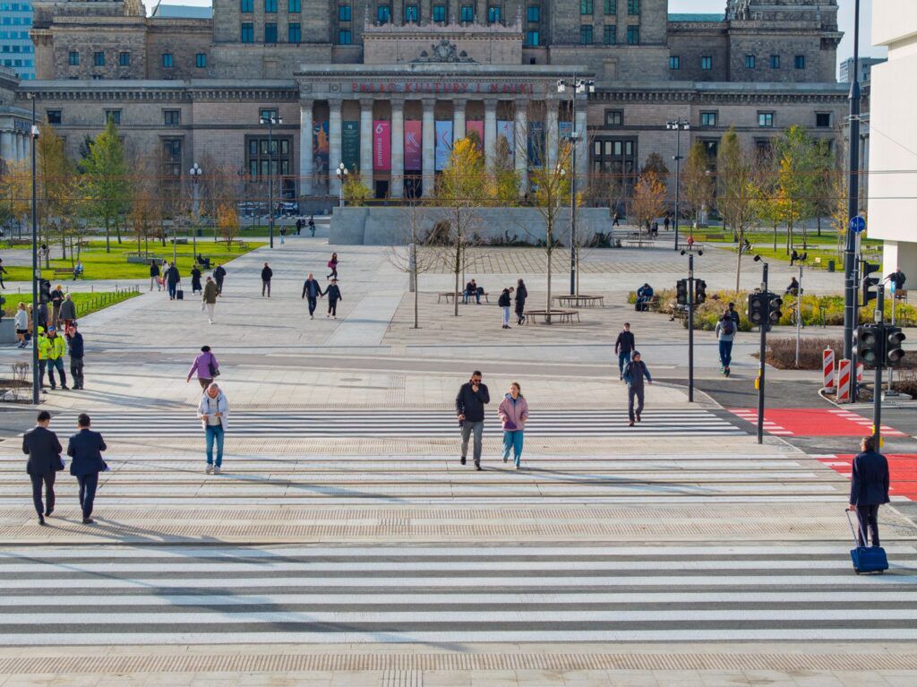 Grupa ludzi przechodzi przez pasy w pobliżu nowoczesnego budynku, w tle widać zieleń i ławki.