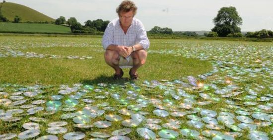 Artist Bruce Munro with his 600,000 CD art installation at Long Knoll, Wiltshire. See SWNS story SWCD: A innovative artist has created a glittering ocean 40 miles inland - using 600,000 recycled COMPACT DISCS. Renowned light installation artist Bruce Munro, 51, created the enormous CD wave to give a feeling of ''reflection'' using commonplace materials. He directed a team of 140 volunteers as they placed the myriad shiny discs in an intricate wave pattern on the grounds of his farmhouse in Kilmington, Wilts., to create 'CD Sea'. The reflective discs are consistently laid out over 10,000 square metres of grass, interrupted only by a single serpentine wave of grass running across the field. Father-of-four Bruce said: ''Lots of the pieces that I work on are about reflection - about what might be and what has gone before.