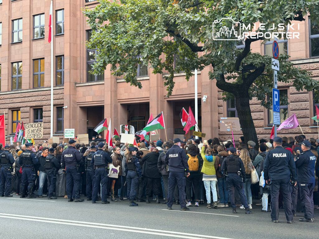 Tłum demonstrantów z transparentami i flagami zgromadzony przed budynkiem, w otoczeniu policji.