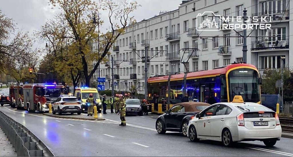 Na ulicy zablokowanej przez pojazdy ratunkowe, strażacy i ratownicy medyczni udzielają pomocy poszkodowanym w wypadku.
