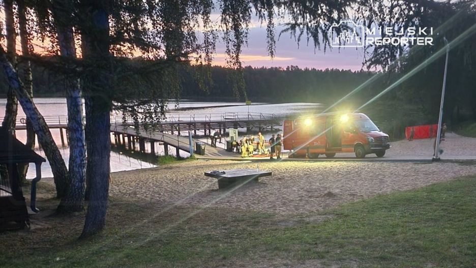 Grupa ratowników pracuje przy brzegu jeziora, obok karetki z włączonymi światłami. W tle widać pomost.