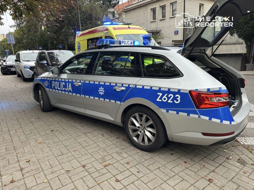 Radiowóz policyjny z niebieskimi światłami zatrzymany na chodniku, obok stoi karetka pogotowia.
