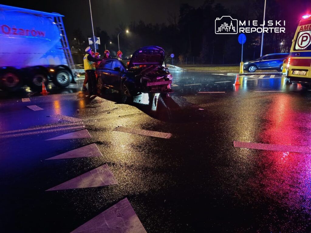 Ratownicy medyczni udzielają pomocy poszkodowanemu przy rozbitym samochodzie na mokrej drodze w nocy.