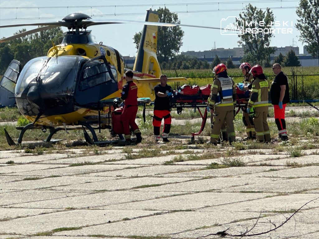 Ratownicy medyczni i strażacy pracują przy helikopterze, przygotowując nosze do transportu poszkodowanego.