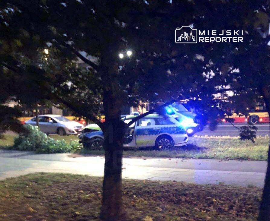 Policyjny radiowóz z włączonymi sygnałami świetlnymi stoi na trawie obok jezdni, a w tle widać inne pojazdy.