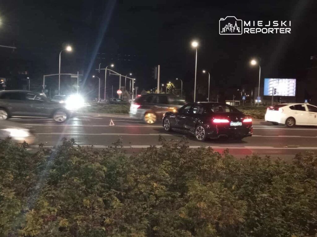 Samochody poruszające się po ruchliwej drodze nocą, z włączonymi światłami, w otoczeniu miejskim.