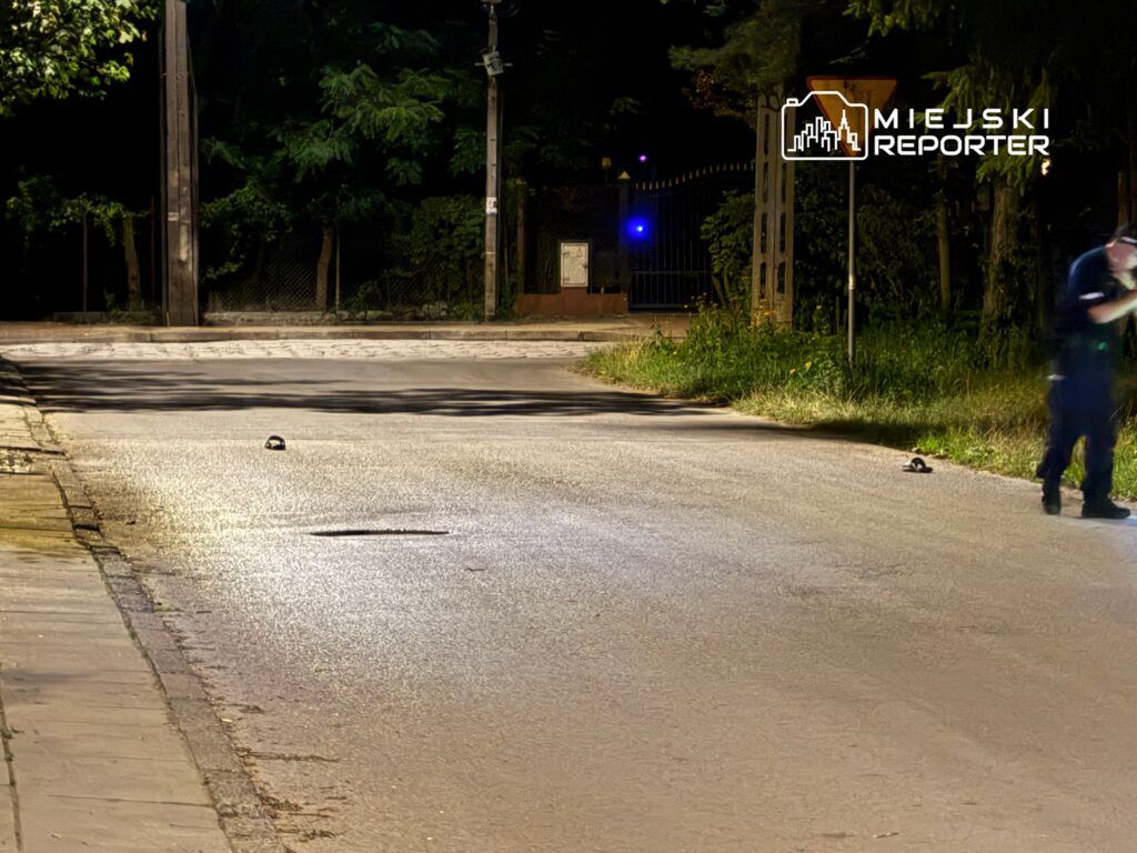 Policjant bada miejsce zdarzenia na oświetlonej ulicy, obok leżą dwa przedmioty na jezdni.