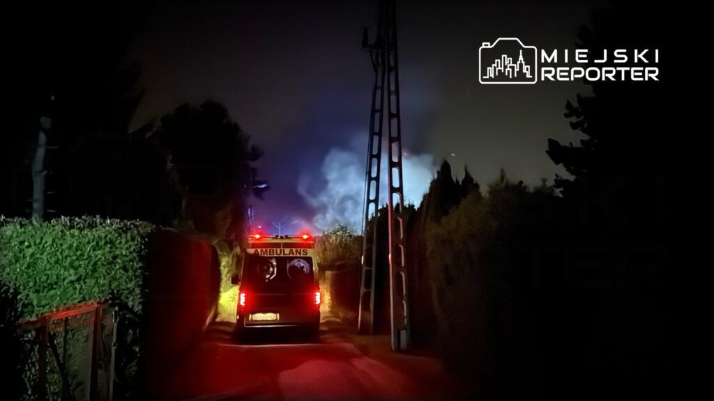 Karetka pogotowia zatrzymana na wąskiej ulicy, w tle niebieskie światła i dym w nocy.