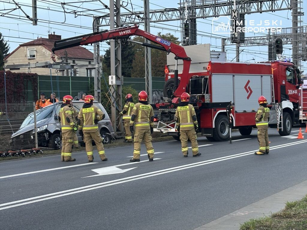 Grupa strażaków w hełmach pracuje przy wypadku drogowym, gdzie samochód osobowy jest podnoszony przez wóz strażacki.