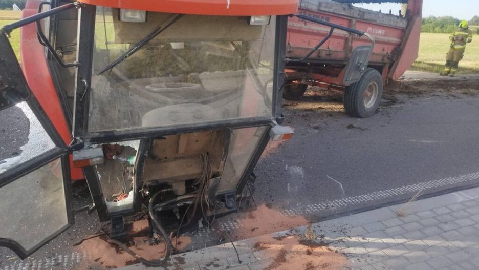 Traktor z uszkodzoną kabiną stoi obok przyczepy, z której wydobywa się błoto, na drodze wiejskiej. W tle widoczny strażak.