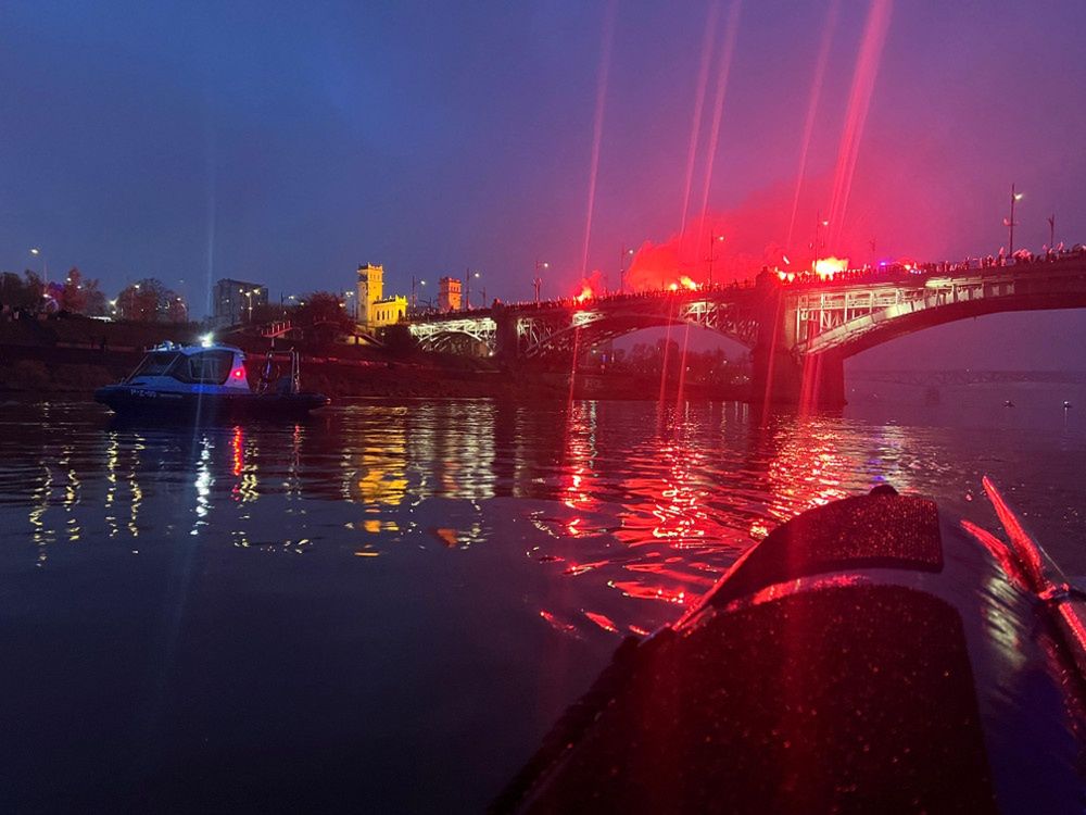 Na rzece widać łódź patrolową, a na moście gromadzą się ludzie z racami, oświetlając nocne niebo.