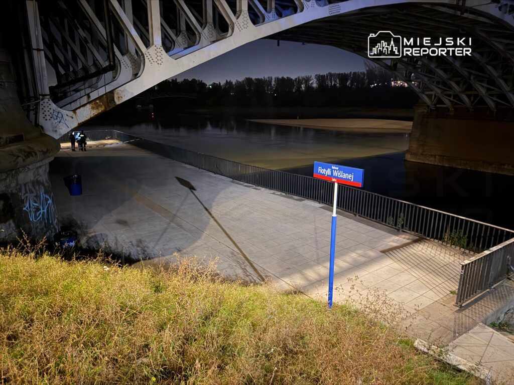 Dwóch ludzi stojących na nabrzeżu pod mostem, obok znaku „Flotyli Wiślanej” w nocnej scenerii.