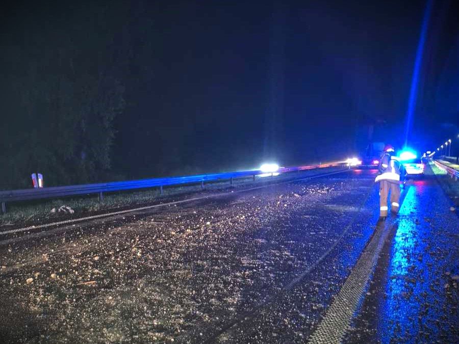 Na drodze w nocy leżą resztki materiałów, a obok stoi strażak w mundurze, podczas akcji ratunkowej.